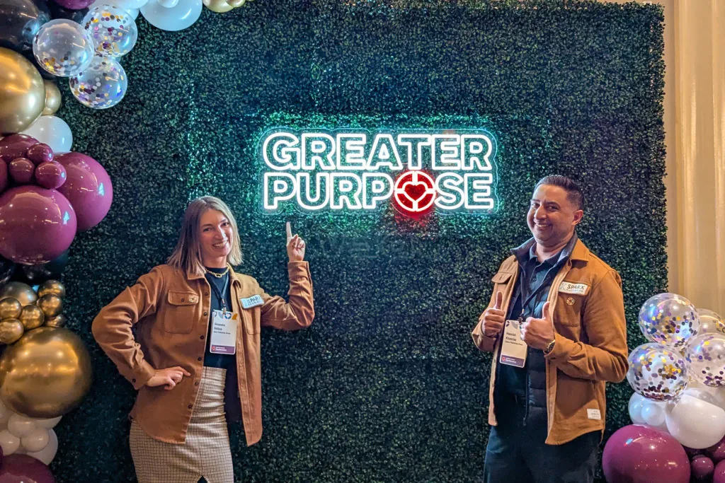 Two people posing in front of a Greater Purpose neon sign framed by balloon garlands