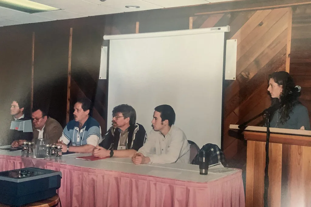 A panel of five people seated at a table with a pink tablecloth beside a projector screen, with a woman speaking at a podium to the right
