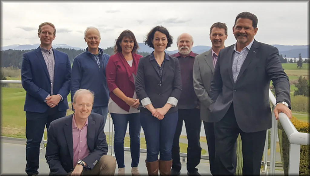 Eight people in business casual attire posing for a group photo on an outdoor balcony overlooking green fields and mountains