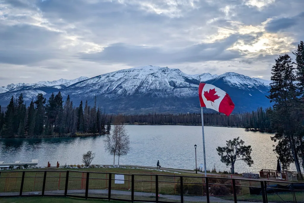 A Canadian flag flying beside a lake with snow-capped mountains and evergreen trees in the background at dusk
