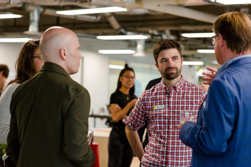 A group of people in an office mingling and talking, one man wearing a name tag reading Graham, with others holding drinks