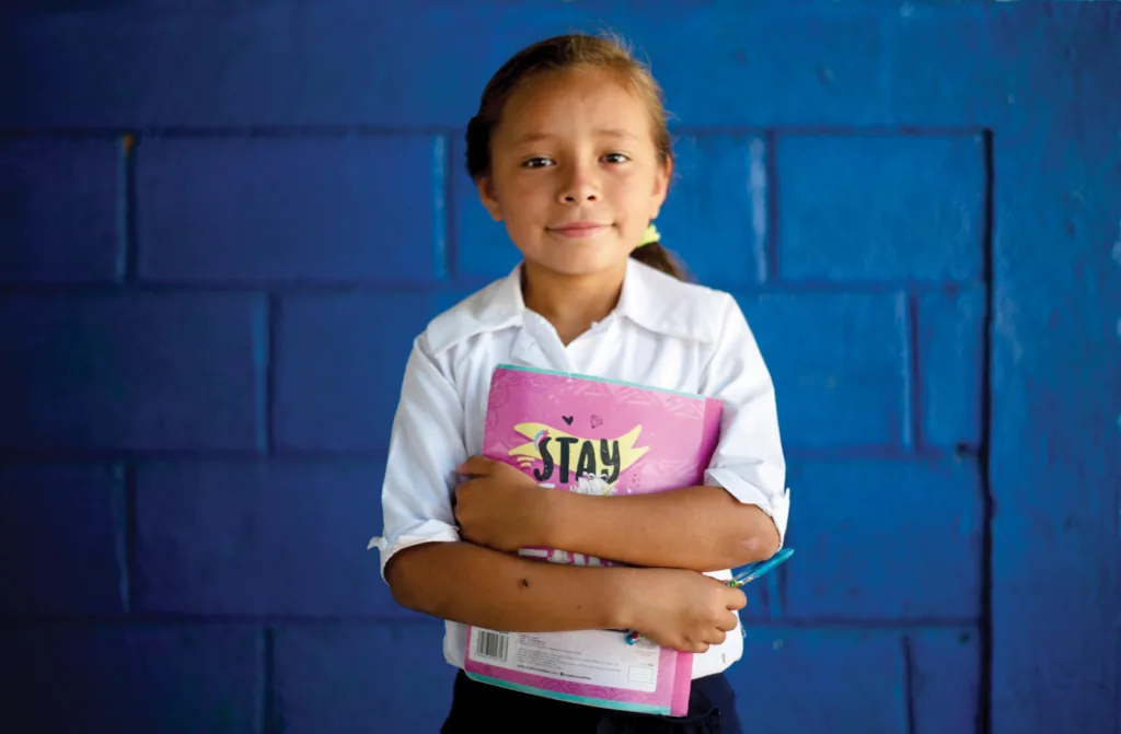 A girl in a white school uniform holding a pink binder against a blue cinder block wall