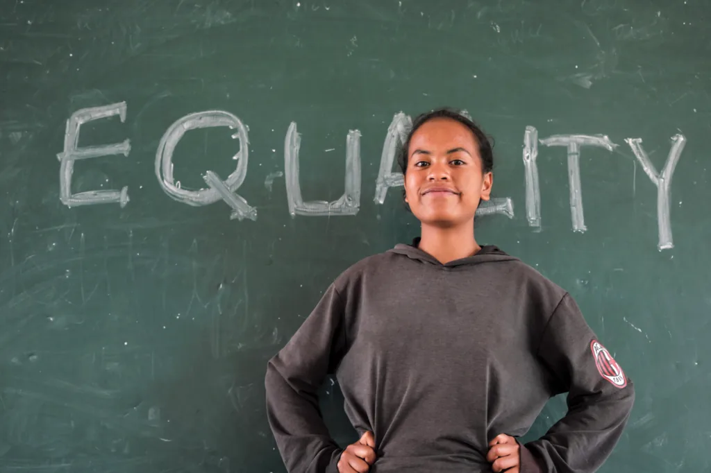A girl standing with hands on her hips in front of a chalkboard with EQUALITY written across it