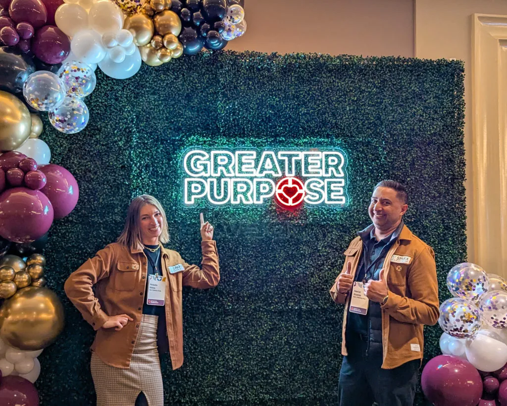 Two people posing in front of a Greater Purpose neon sign framed by balloon garlands