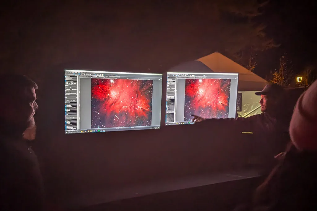 Two men viewing dual computer monitors displaying a red nebula astrophotography image at an outdoor nighttime event