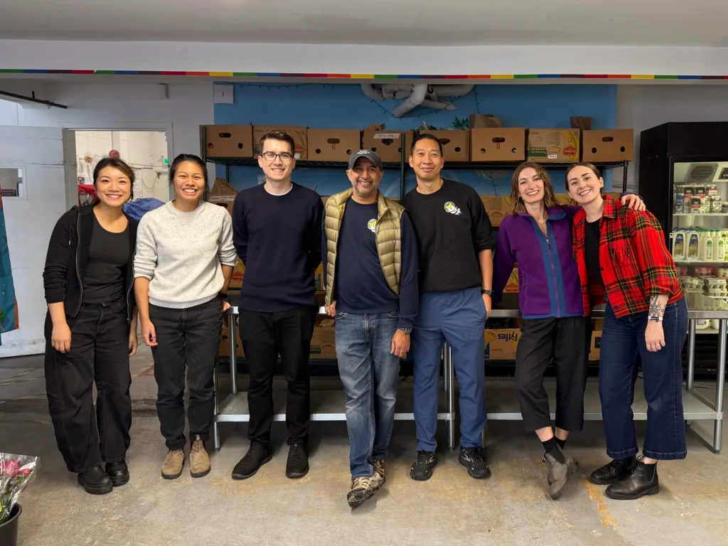 A group of seven people posing together in a food bank warehouse lined with shelved food boxes