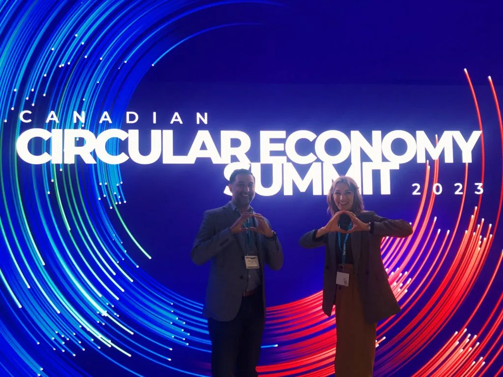 A man and woman forming heart shapes with their hands in front of a Canadian Circular Economy Summit 2023 sign