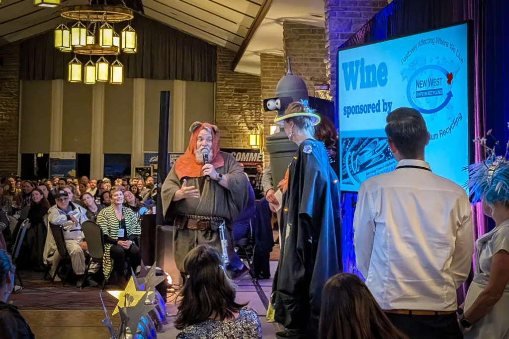 A person in an Ewok-style costume speaking into a microphone on stage at an awards event, with a screen reading Wine sponsored by New West Gypsum Recycling and costumed attendees nearby