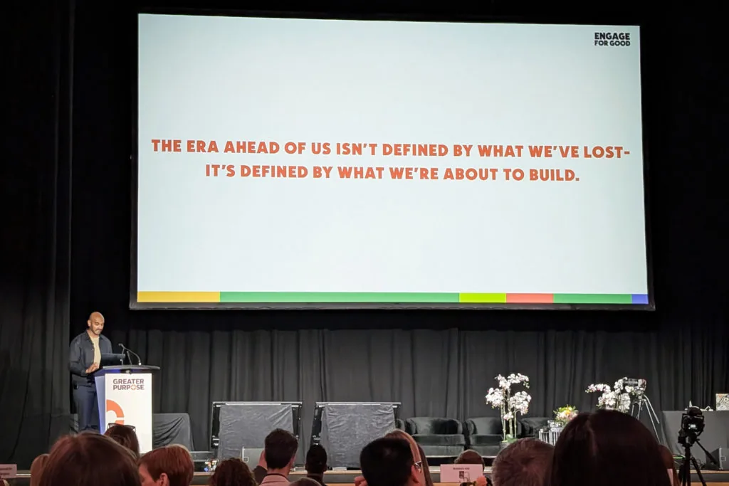 A speaker at a podium below a conference screen reading The era ahead of us isn't defined by what we've lost, it's defined by what we're about to build
