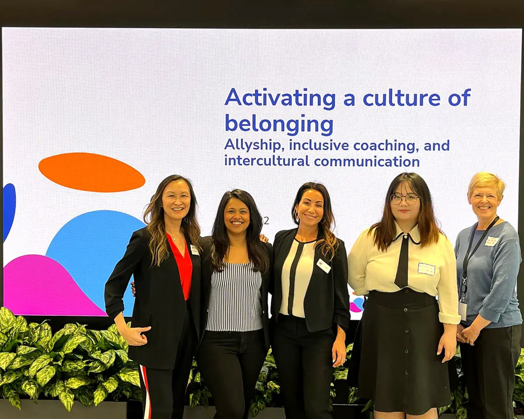 Five women stand together on stage in front of a screen reading Activating a culture of belonging: Allyship, inclusive coaching, and intercultural communication