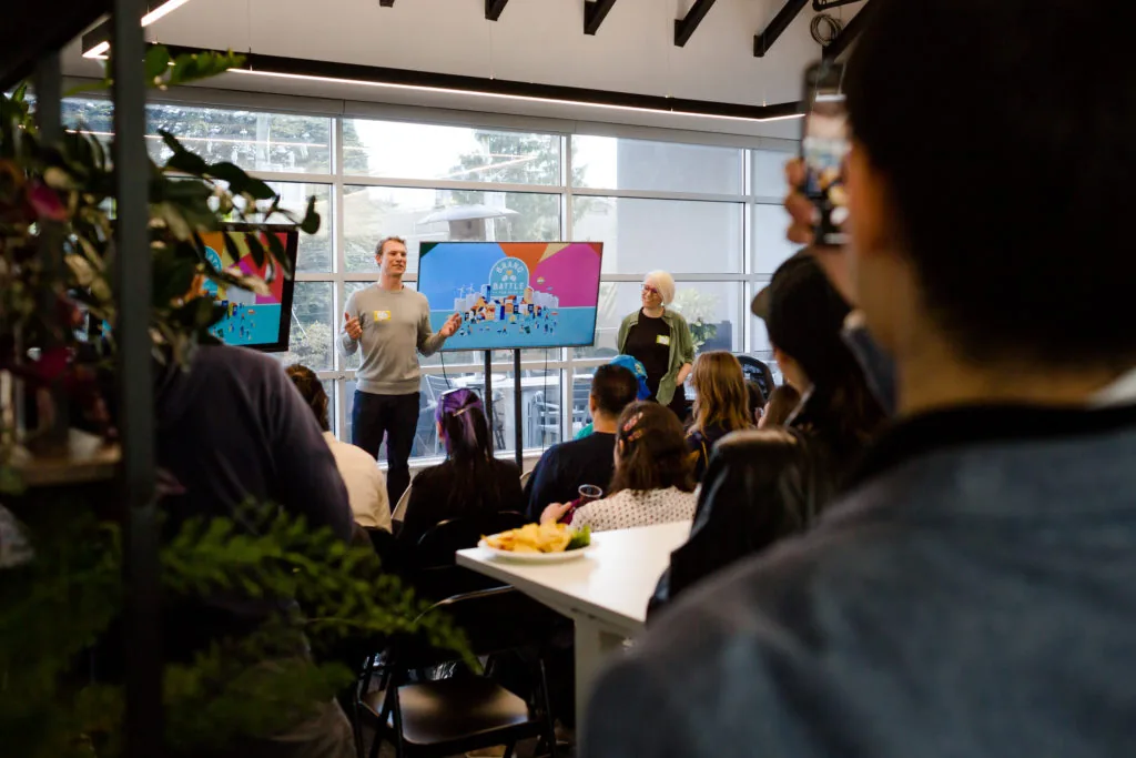 A man presents to a seated audience in front of two screens displaying a colourful Urban Battle graphic, with a woman standing beside him
