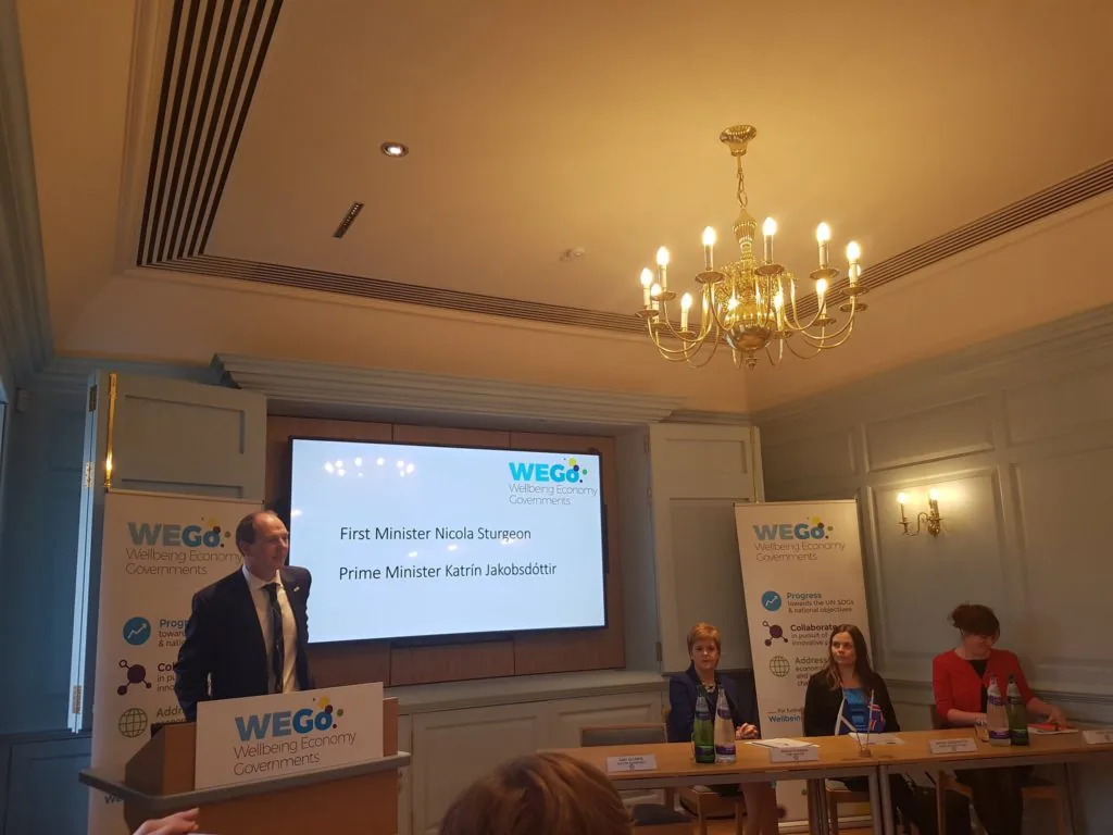 A man speaks at a podium bearing the WEGo Wellbeing Economy Governments logo, beside a screen naming First Minister Nicola Sturgeon and Prime Minister Katrin Jakobsdottir, with three seated panelists at a table