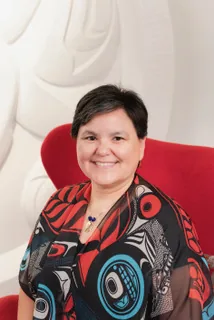 A woman wearing an Indigenous-patterned shawl seated in a red chair against a carved wall relief