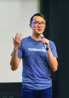 A man in a Moment-branded t-shirt speaks into a microphone while gesturing with his hand
