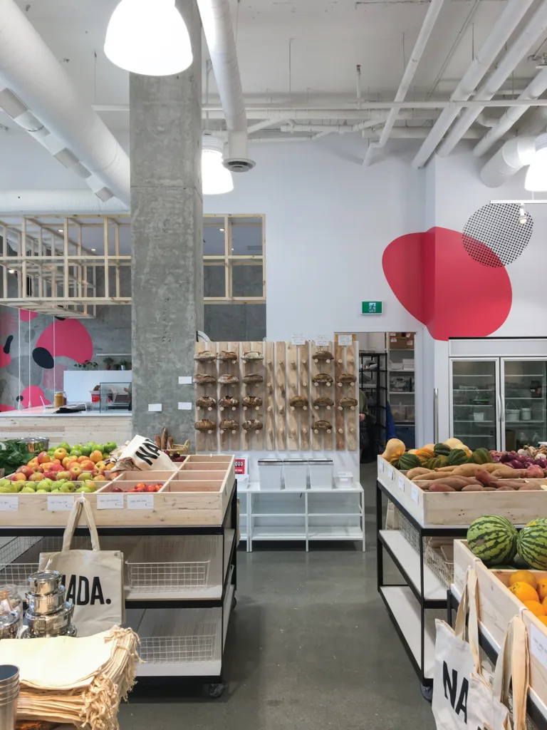 The interior of a grocery store with bare wooden produce bins of fruit and vegetables, a bread display wall, and a pink graphic mural