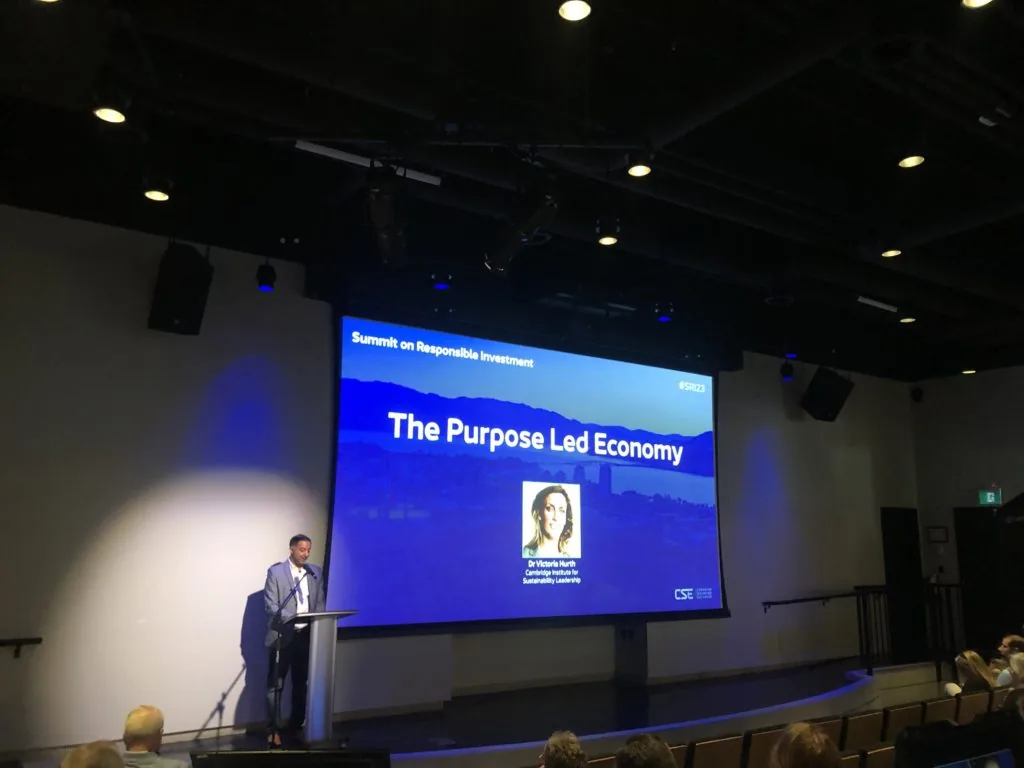 A man speaking at a podium below a screen reading The Purpose Led Economy at the Summit on Responsible Investment