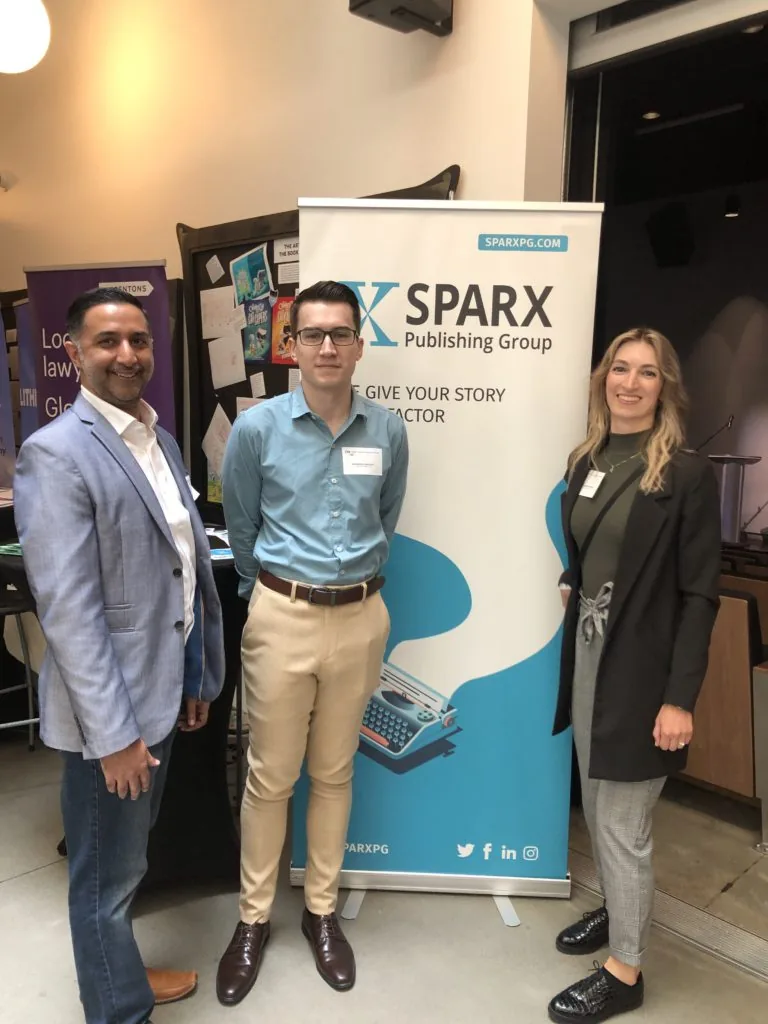 Three people standing beside a Sparx Publishing Group banner reading We Give Your Story the X Factor