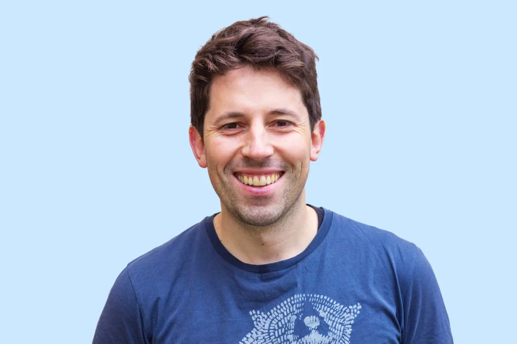 A studio headshot of a smiling man in a blue t-shirt against a light blue background