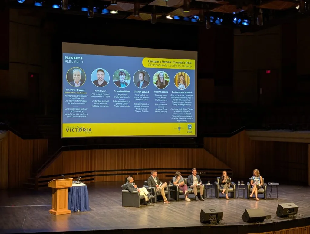 A conference auditorium stage with six panelists seated below a large screen reading Plenary 3, Climate x Health: Canada's Role