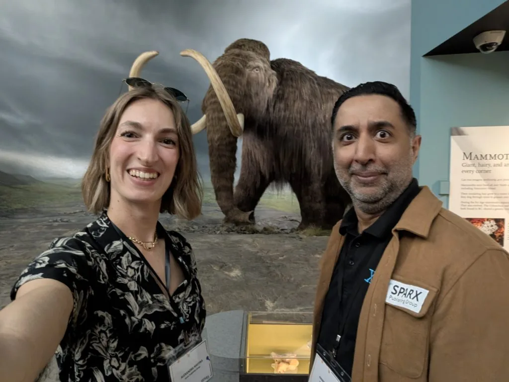 A man and woman pose for a selfie inside a museum exhibit, with a life-size woolly mammoth diorama behind them