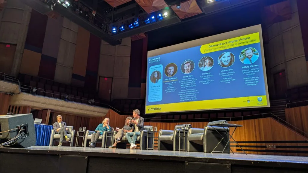 A conference stage with four panelists seated below a large screen reading Plenary 8, Democracy's Digital Future
