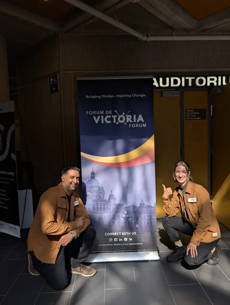 A man and woman kneeling on either side of a Forum de Victoria Forum banner reading Bridging Divides, Inspiring Change outside an auditorium door