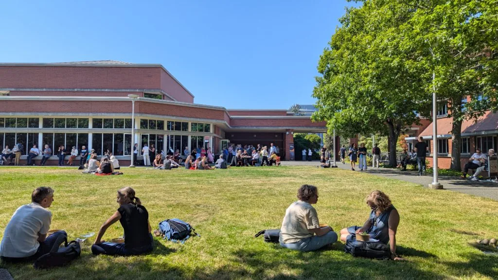 Attendees sitting on a grassy lawn in small groups outside a brick conference building on a sunny day