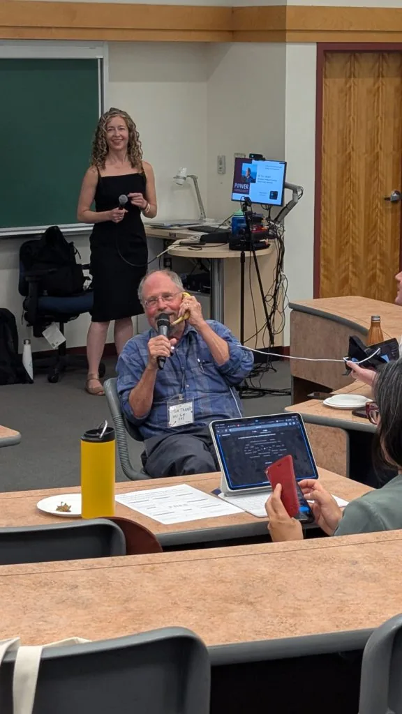 An older man speaking into a microphone while holding a banana to his ear like a phone, with a woman standing beside a chalkboard behind him