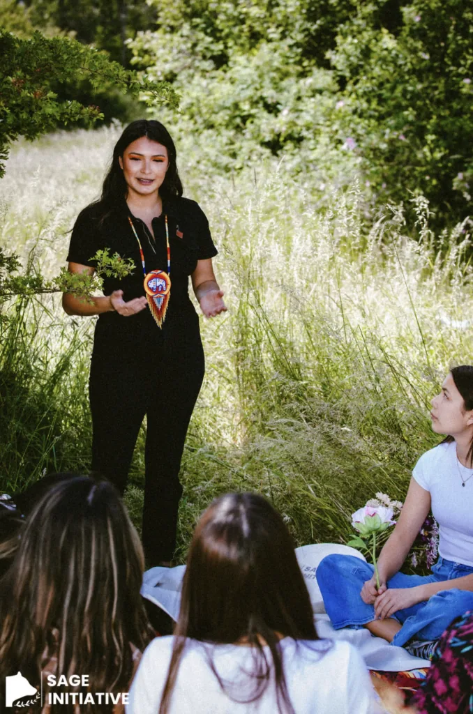 A woman wearing a beaded Indigenous pendant necklace speaks to a seated group outdoors in tall grass, with a Sage Initiative logo overlaid
