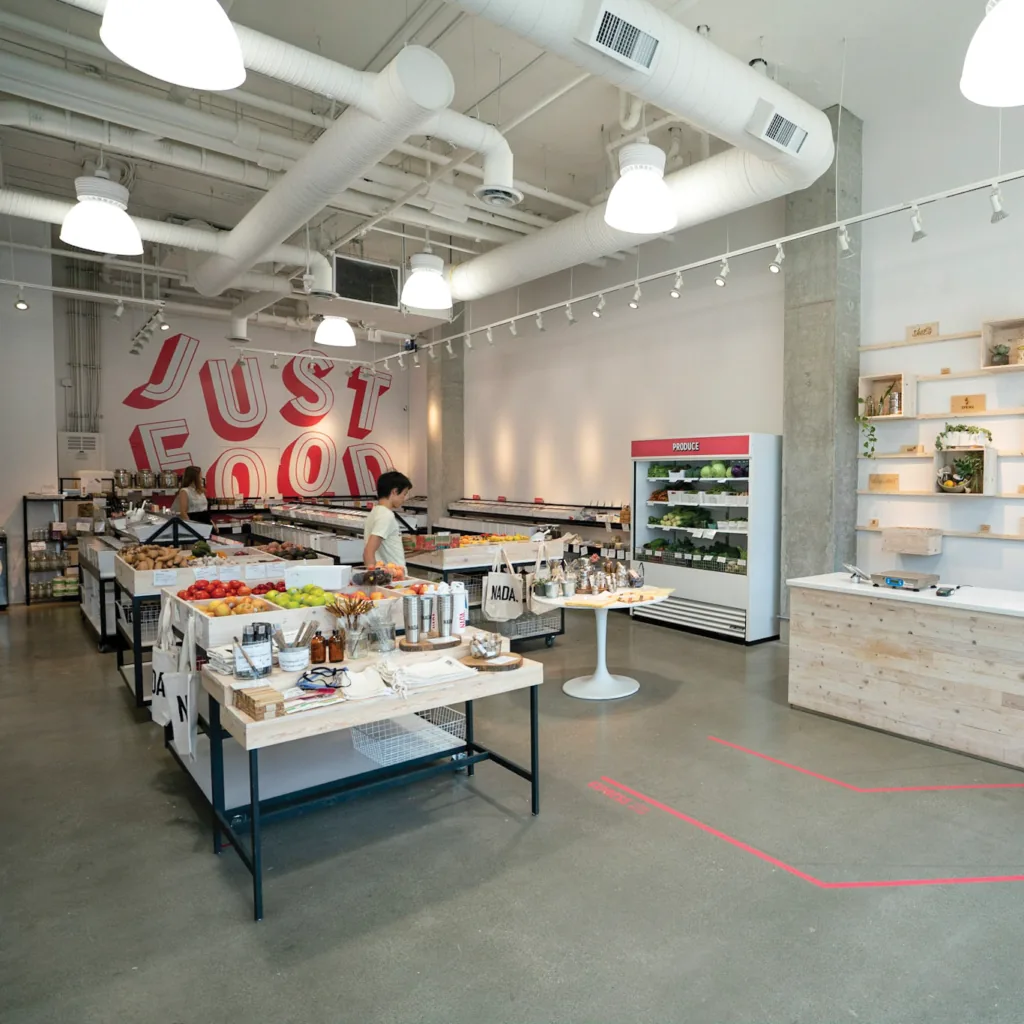 The interior of a grocery store with a wall reading Just Food, produce tables, a chilled produce case, and a checkout counter
