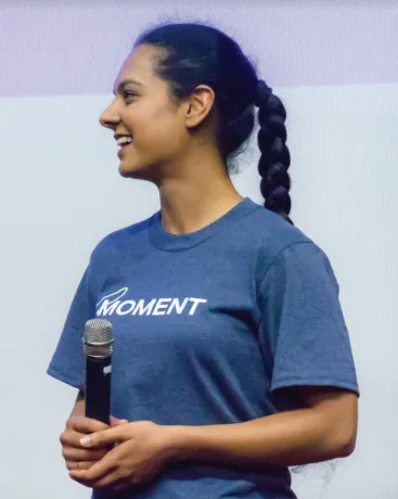 A woman in a Moment-branded t-shirt stands in profile holding a microphone, smiling