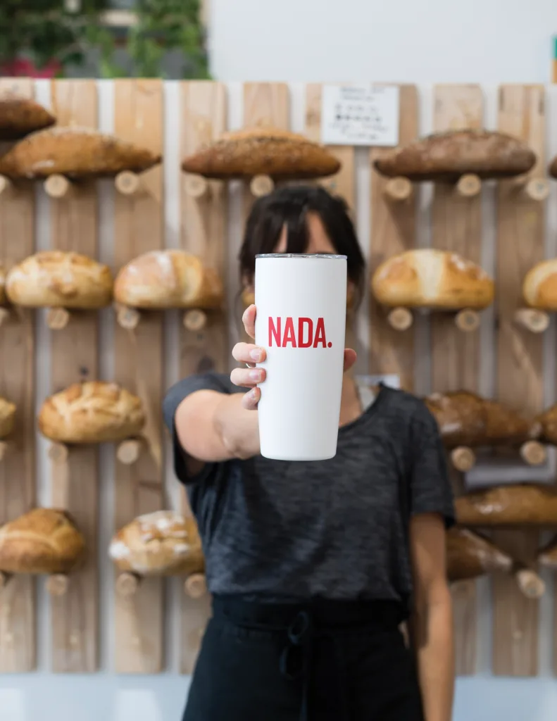 A person holds up a white NADA-branded tumbler in front of a wall of loaves of bread displayed on wooden pegs