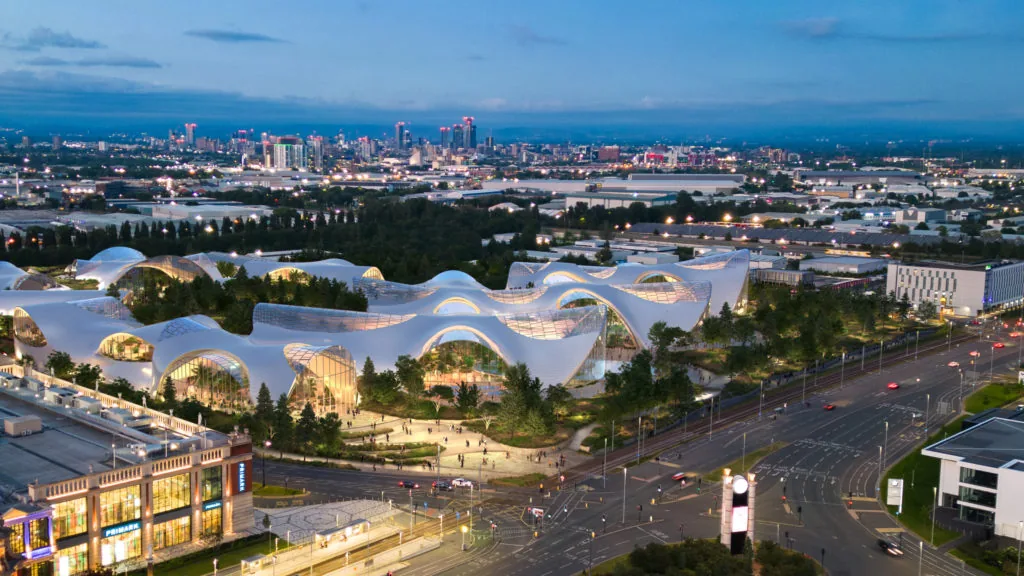 An aerial rendering of the Therme Manchester wellbeing complex, its curved glass roofline lit at dusk beside a highway