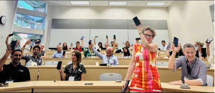 A crowd of seated conference attendees in a lecture hall holding up their phones, with a woman in a colourful dress standing at the front