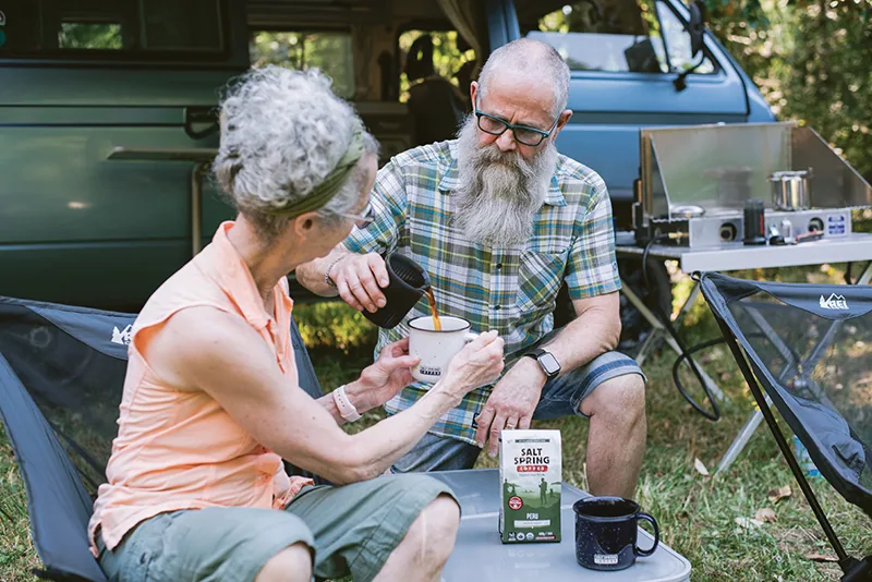 An older couple seated at a campsite beside a van, the man pouring coffee into a mug held by the woman, with a Salt Spring Coffee bag on the table