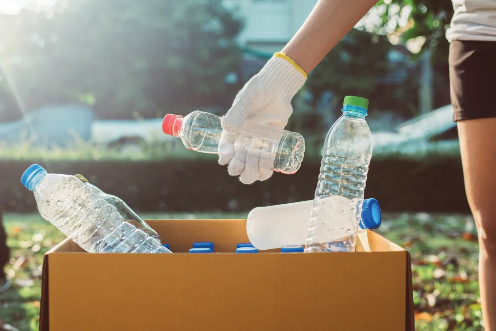 A gloved hand placing a plastic water bottle into a cardboard box already holding several empty bottles