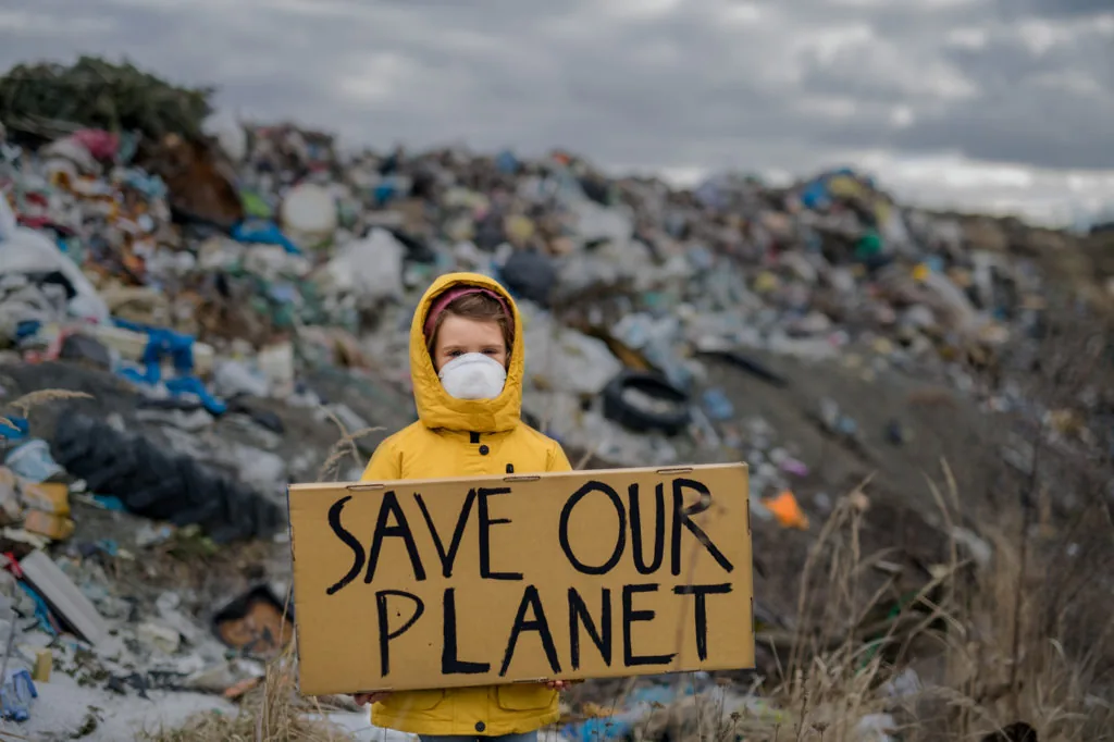 A child in a yellow raincoat and face mask holding a cardboard sign reading Save Our Planet in front of a large landfill